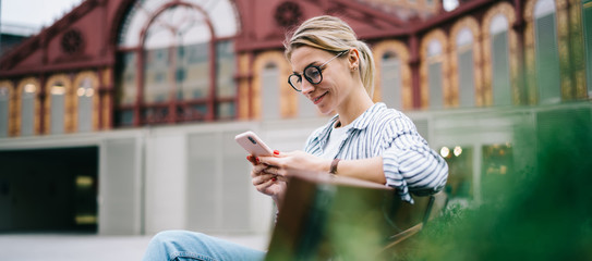 Smiling woman in eyeglasses using smartphone on street bench