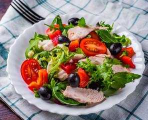 Fresh salad with chicken breast, arugula, black olives,red pepper, lettuce, fresh sald leaves and tomato on a white plate on wooden table background