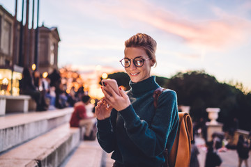 Positive young hipster girl typing text message using 4g wireless for networking and online...