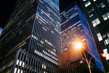 Evening view of modern constructed futuristic buildings with shining exterior lights located in megalopolis center,tall skyscrapers with luxury apartments or business offices for rent in downtown.