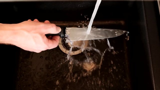 Wash the knife. A knife in a spray of water. Male hand washes a sharp knife under the stream of water in a black sink