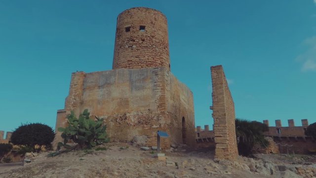 A slow motion low angle view of an old watchtower of castle capdepera with clear teal sky.