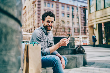 Naklejka premium Portrait of happy bearded hipster blogger smiling at camera while chatting online on smartphone using 4G internet sitting outdoors near mall with paper bags after great shopping in black friday