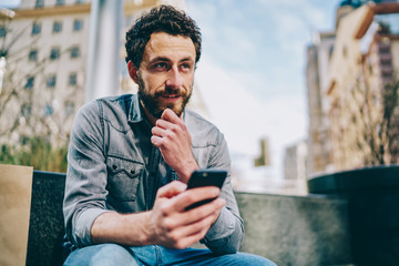 Bearded hipster guy sitting in urban setting and thinking on publication in own internet blog using...