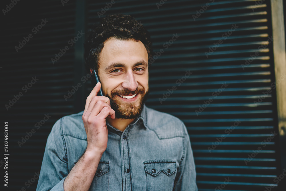Poster positive bearded hipster guy with beard smiling at camera while communicating with best friend on sm
