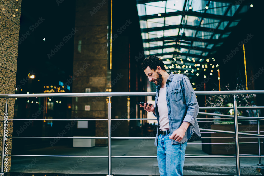 Poster side view of casual dressed hipster guy strolling in urban setting near business building and readin