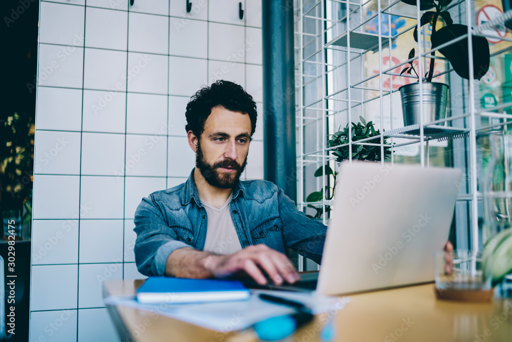 Poster pensive bearded young man working on freelance on development of website at modern laptop using wire