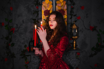 Fototapeta premium A beautiful girl in a magnificent, red rococo dress holds in her hands a candlestick with candles standing in the dark, against the background of the fireplace, window, and flowers.