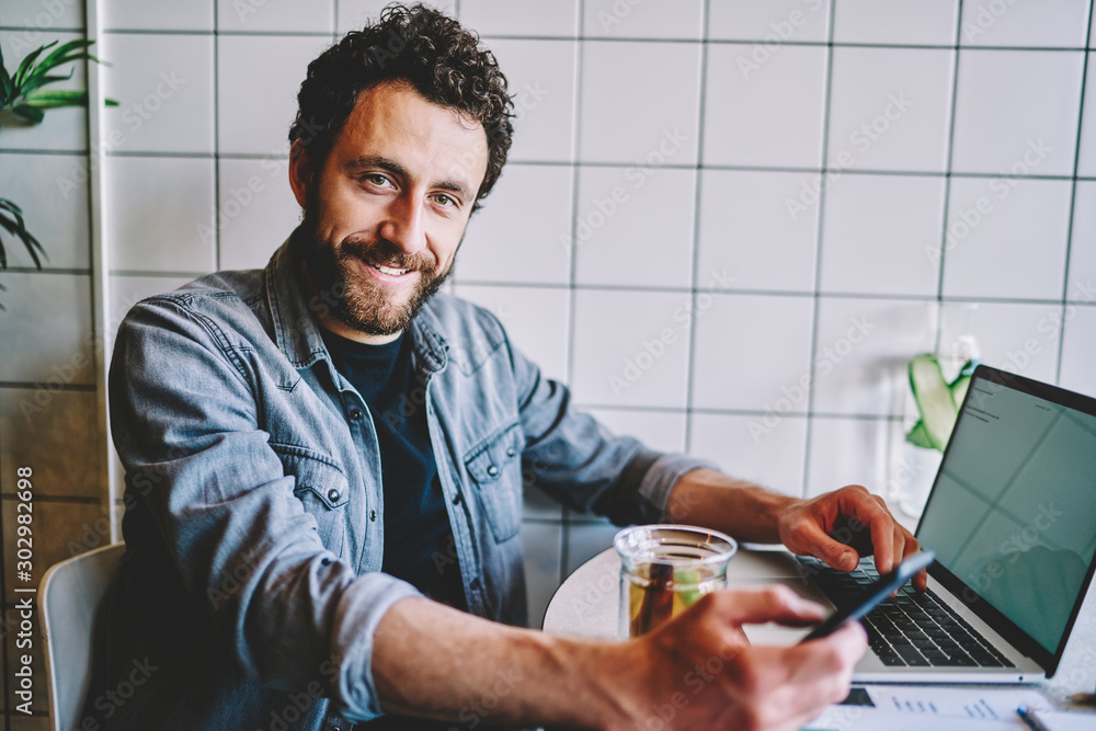 Poster portrait of bearded positive hipster guy smiling at camera while sharing media from smartphone on mo