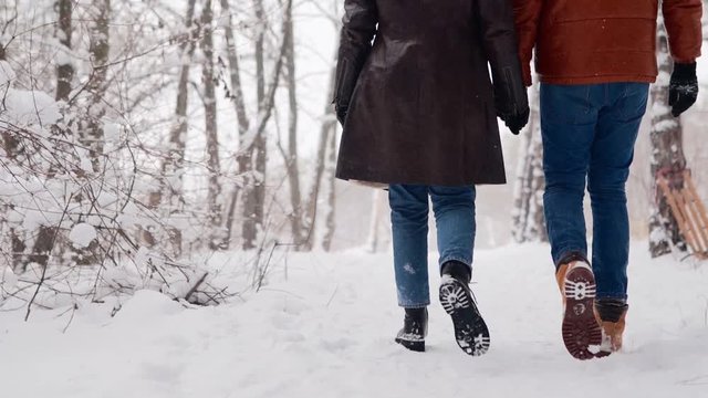 Back view of loving couple feet walking in a park on snowfall. Man and his girlfriend holding hands enjoying snow on winter day. Hipster woman stroll in snowy forest with boyfriend in stylish clothing