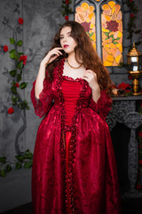 Beautiful girl in a magnificent, red rococo dress. Against the background of the fireplace, window, and flowers.