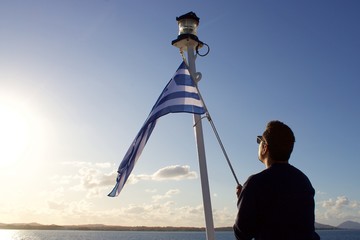Silhouette of a sailor man raising a Greek flag on a pile of a beacon at the stern of a liner ferry...