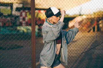 Young attractive woman in jeans jacket, shorts, red top and trucker hat posing over metal fence and...
