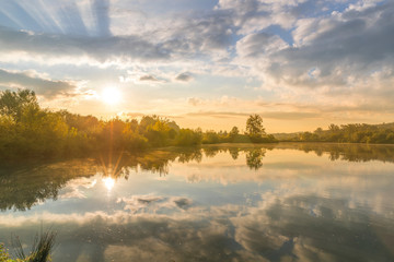 Pond at the morning sunrise over trees lying near water and moving clouds over water with reflection on water surface.