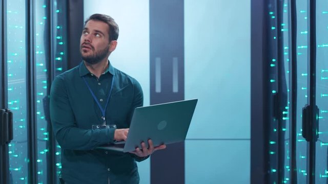 Bearded system administrator working on notebook inspecting operating database server rack cabinets in big modern data center. Cyber security and technology concept.