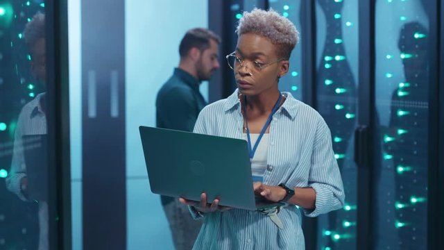 Black female IT engineer upgrading hardware in rack server room corridor working on computer laptop cooperating with male specialist at big data center.