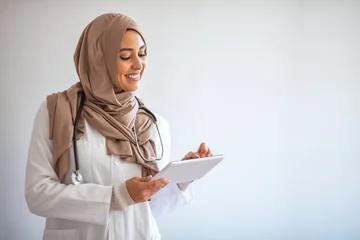 Fotobehang Muslimah doctor with stethoscope holding a tablet computer isolated in grey background. Confident Muslim medical student pose at hospital. Confident Muslim female doctor standing inside hospital © Dragana Gordic