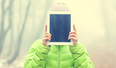 Millennials girl holding the tablet as the face - blank screen with copy space - magical misty forest in the background - girl wearing a down jacket i prezętuje blank screen on tablet