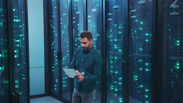 IT professional with digital tablet monitoring system performance of database network servers working on rack in high tech data center. Webhosting, cloud computing.