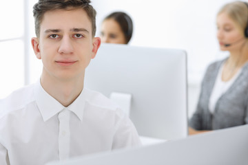 Fototapeta premium Call center. Group of operators at work. Focus at young guy receptionist in headset at customer service white colored office. Business concept