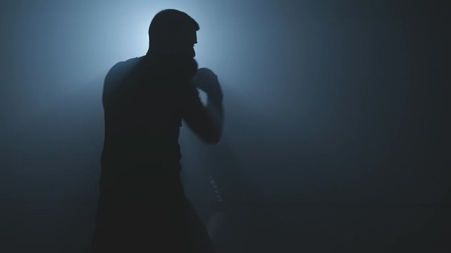 Male boxer training on black smoky background with light. Shadow fight. Silhouette of boxer preparing for big fight. Strength and motivation. ide view. Close-up shot in 4K, UHD