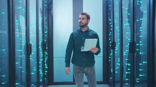 Caucasian IT administrator walking in database corridor inspecting operating rack servers network doing diagnostics on digital laptop in big data center.