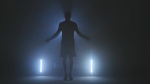 Boxer jumping using jump rope while warming in dark smoky gym in slow motion. Silhouette of young boxer skipping. Full length in 4K, UHD