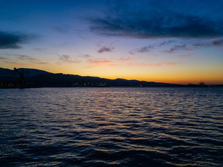 Beautiful colorful sunrise over the surface of the water of the sea with small waves with a mountainous contour of the shore of a coastal town against a blue cloudy sky.