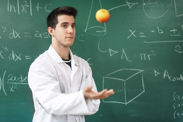 Smiling student with white coat throwing an apple up, on green chalkboard with equations background...