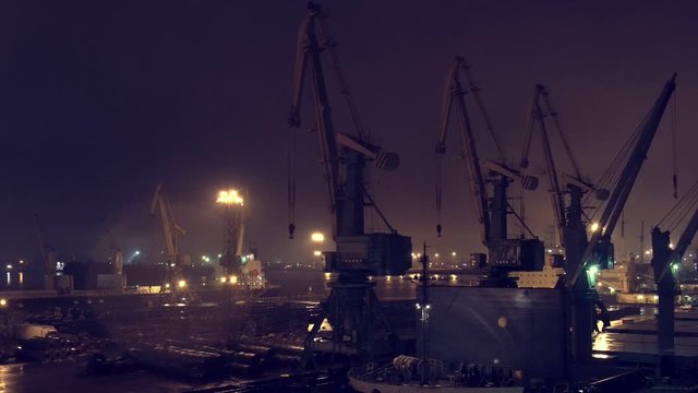 Ship leaving harbor sailing along the jetty pier with many crane spreaders and containers at the night