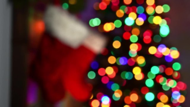 An out of focus Christmas theme background with multi-colored twinkle lights on a Christmas tree as the stocking are hung by the fireplace with a warm glow of a fire at night