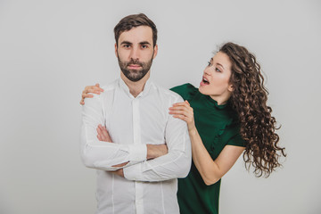 Serious man is standing, looking straight forward, his hands folded, woman looking at him from behind his back, confused.