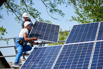 Two workers installing heavy solar photo voltaic panel on tall steel platform on green tree and...