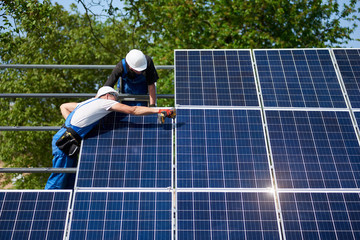 Two technicians working with electrical screwdriver installing shiny solar photo voltaic panel to metal platform system on green tree thick foliage background. Green energy production concept.