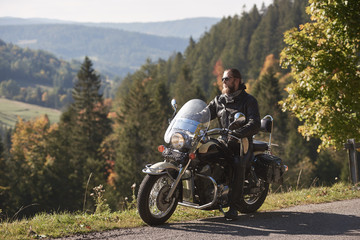 Fototapeta na wymiar Handsome bearded biker in black leather jacket and sunglasses sitting on modern motorcycle on country roadside, on blurred background of foggy green hills covered with dense spruce forest.