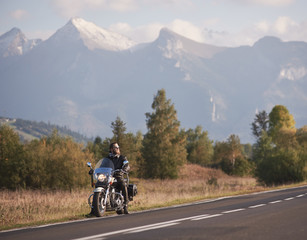 Young bearded biker in black leather jacket and sunglasses sitting on bike on country roadside on blurred background of green rural landscape, distant white sunny mountain peaks and bright sky.