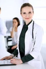 Doctor woman at work with patient and colleague at background. Physician filling up medical documents or prescription while standing in hospital reception desk. Data in medicine and health care