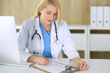 Doctor woman at work while sitting at the desk in hospital or clinic. Blonde cheerful physician filling up medical records form. Data and best service in medicine and healthcare