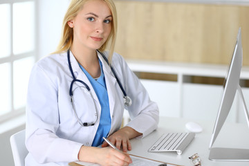 Doctor woman at work while sitting at the desk in hospital or clinic. Blonde cheerful physician filling up medical records form. Data and best service in medicine and healthcare