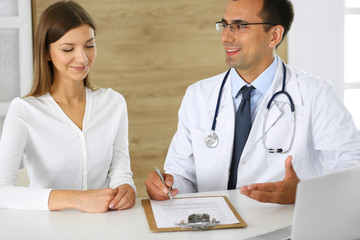 Doctor and patient discussing the results of a physical examination while sitting at a desk in a clinic. A male doctor using a clipboard to fill out a medical history of a young woman's medication