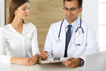 Doctor and patient discussing the results of a physical examination while sitting at a desk in a clinic. A male doctor using a clipboard to fill out a medical history of a young woman's medication