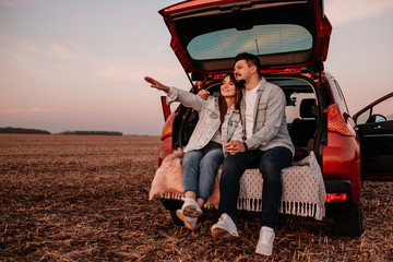 Young Happy Couple Dressed Alike in White Shirt and Jeans Sitting at Their New Car Trunk, Beautiful...