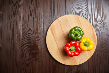 Red, green, and yellow bell peppers, cutting board on wooden background with copy space. Three sweet peppers in different colors on brown table, vegetable ingredient, healthy food, top view