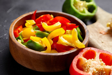 Chopped bell peppers in wooden bowl on black background. Red, green, yellow peppers. Colorful pieces of sweet peppers. Vegetable salad ingredient, cooking healthy diet food