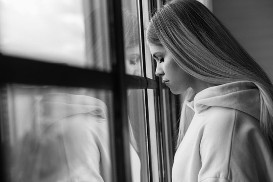 Black and white picture of young upset woman in stress. Stand at window and keep eyes closed. Alone in room. Depression and sadness. Side view.