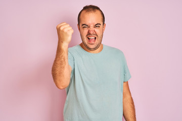 Fototapeta premium Young man wearing blue casual t-shirt standing over isolated pink background angry and mad raising fist frustrated and furious while shouting with anger. Rage and aggressive concept.