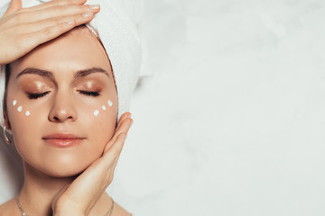 Cut view of young woman's face with clear skin. Model isolated over grey background. Touching face with hands. White cream spots on cheekbones. Beauty daily routine.