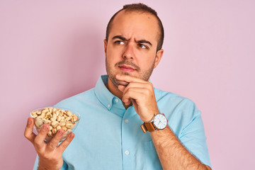 Young man holding bowl with pistachios standing over isolated pink background serious face thinking...