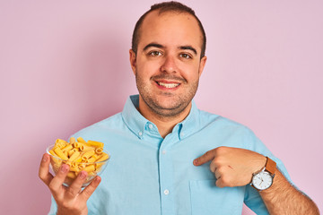 Young man holding bowl with macaroni pasta standing over isolated pink background with surprise face pointing finger to himself