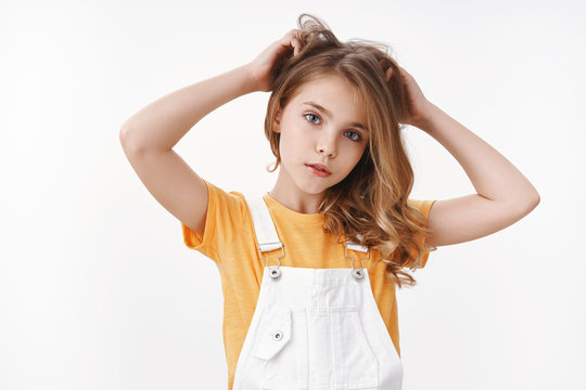 Tender lovely young blond girl in summer dungarees over yellow t-shirt, look thoughtful and cute camera, scratch hair bothered, touch hairstyle, suffer head lice, lacking vitamins, white background